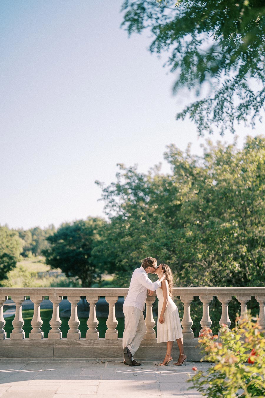 Rachel and Matt kiss beside a stone terrace railing with trees and gardens behind them.