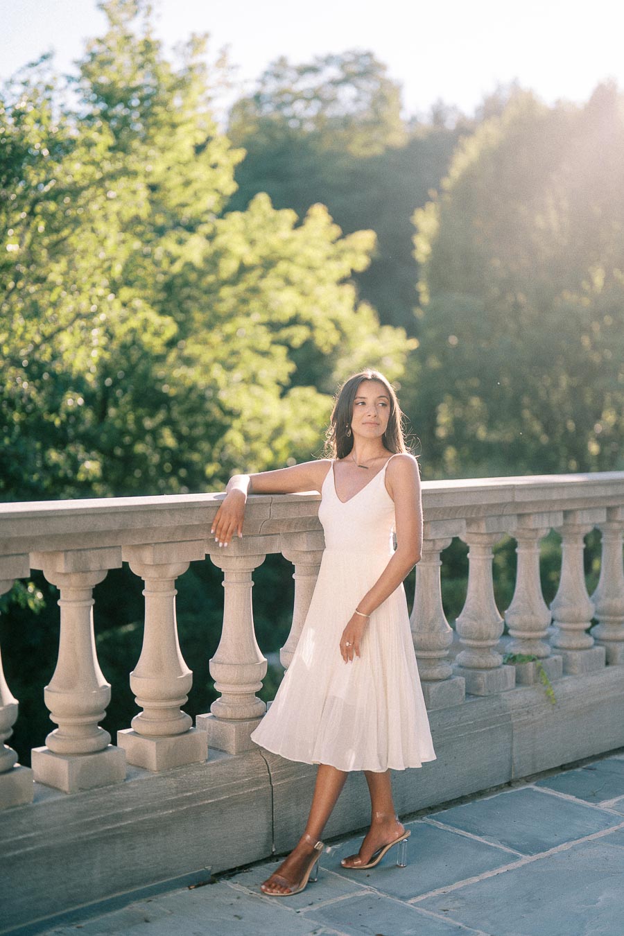 Rachel stands by a stone balustrade in a white dress, with trees and warm sunlight behind her.