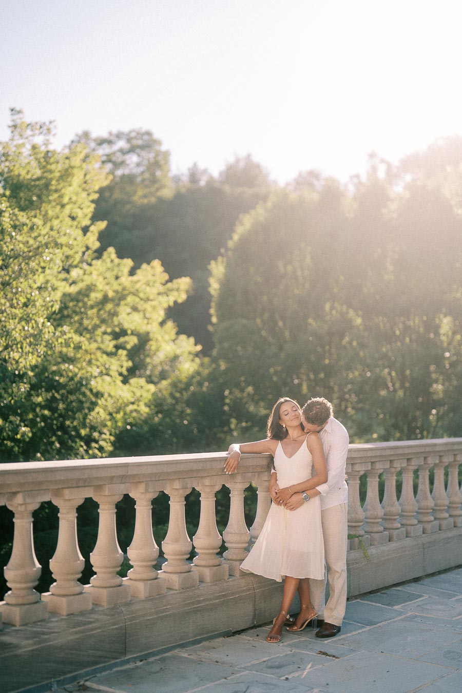 Rachel leans against a stone balustrade while Matt embraces her from behind in warm evening light.