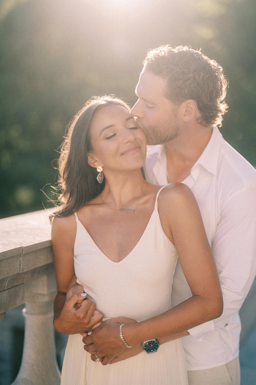 Matt kisses Rachel’s temple as they embrace by a stone railing in soft evening light.