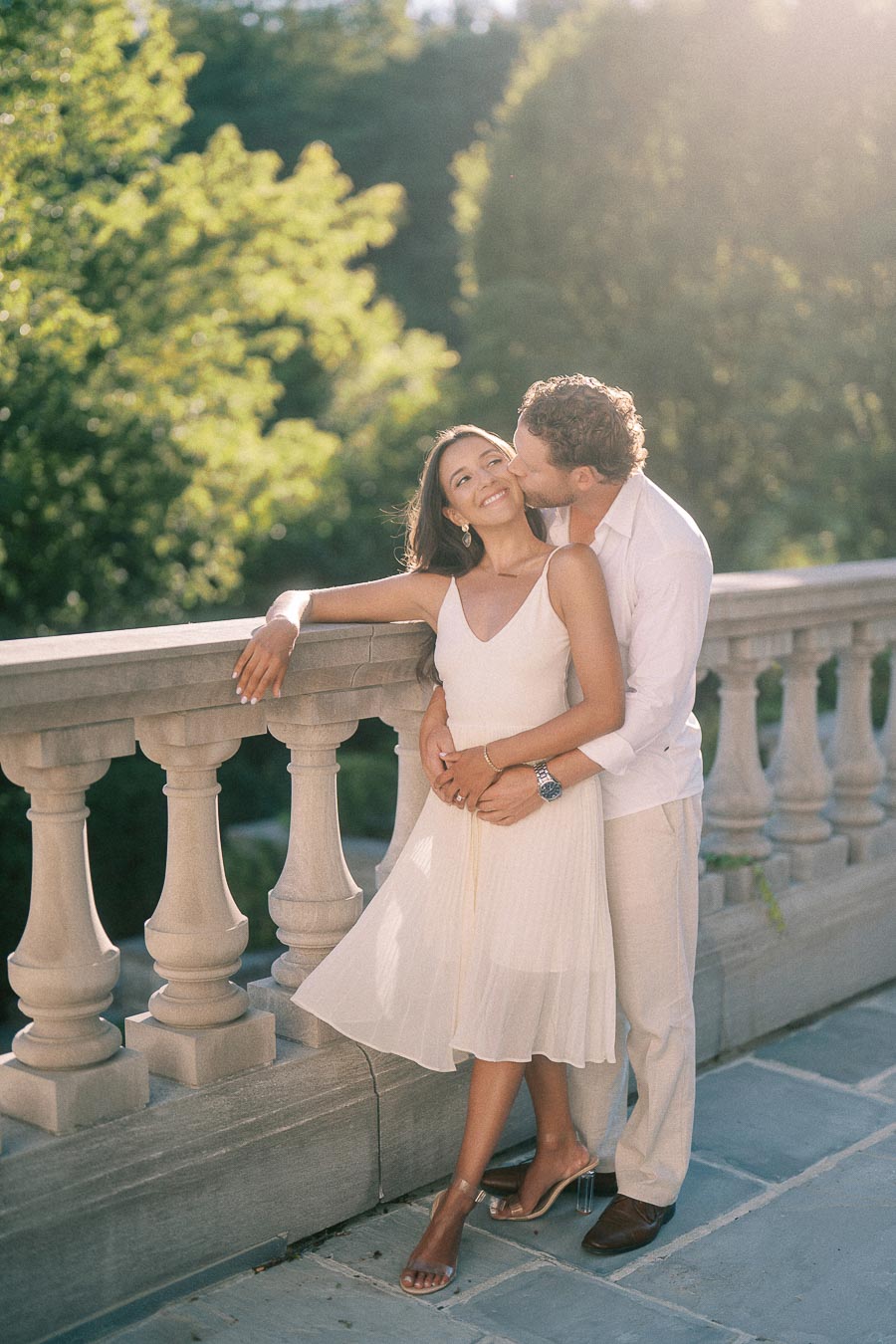 Matt kisses Rachel on the cheek as they stand by a stone balustrade in warm sunlight.