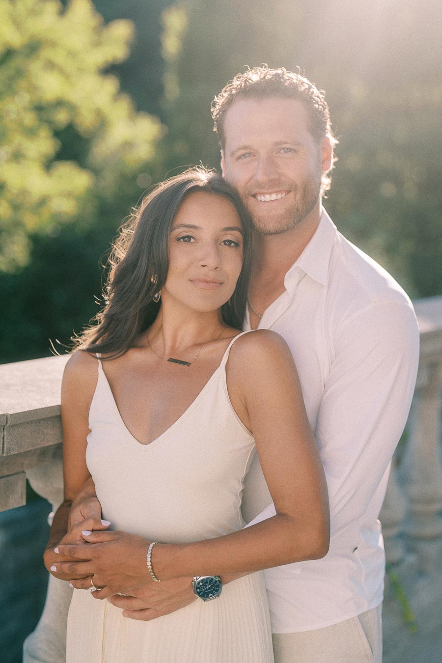 Rachel and Matt stand closely together by a stone railing in warm sunlight with trees behind them.