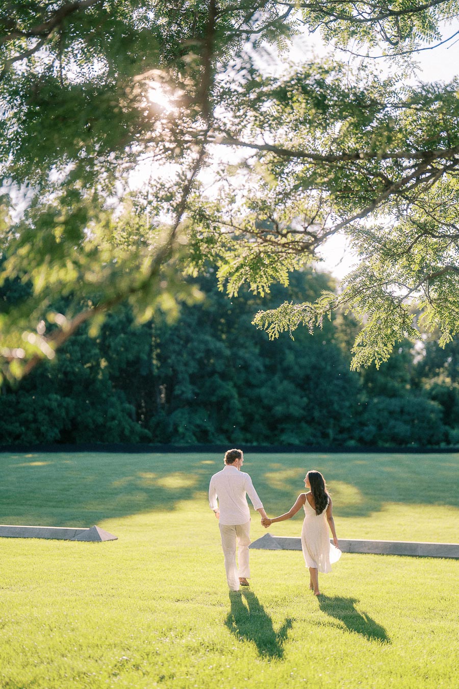 Rachel and Matt hold hands while walking across a sunlit lawn beneath leafy trees in Aurora, Ontario.