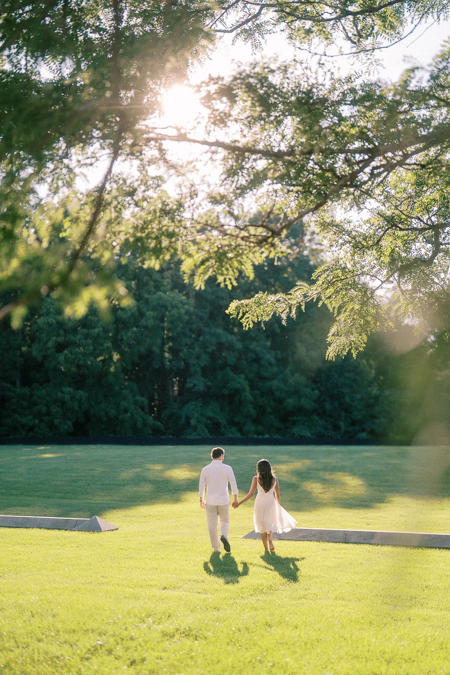 Rachel and Matt walk hand in hand across a sunlit lawn beneath leafy trees in Aurora, Ontario.