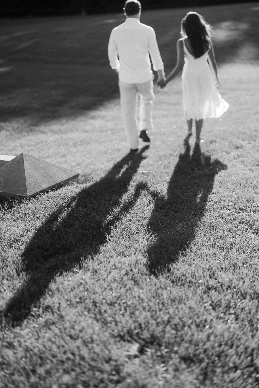 Rachel and Matt walk hand in hand across a sunlit lawn, their long shadows stretching over the grass.