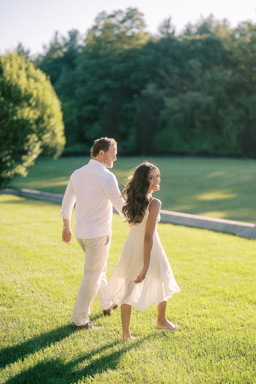 Rachel and Matt walk hand in hand across a sunny grass lawn in Aurora, Ontario, looking back toward the camera.