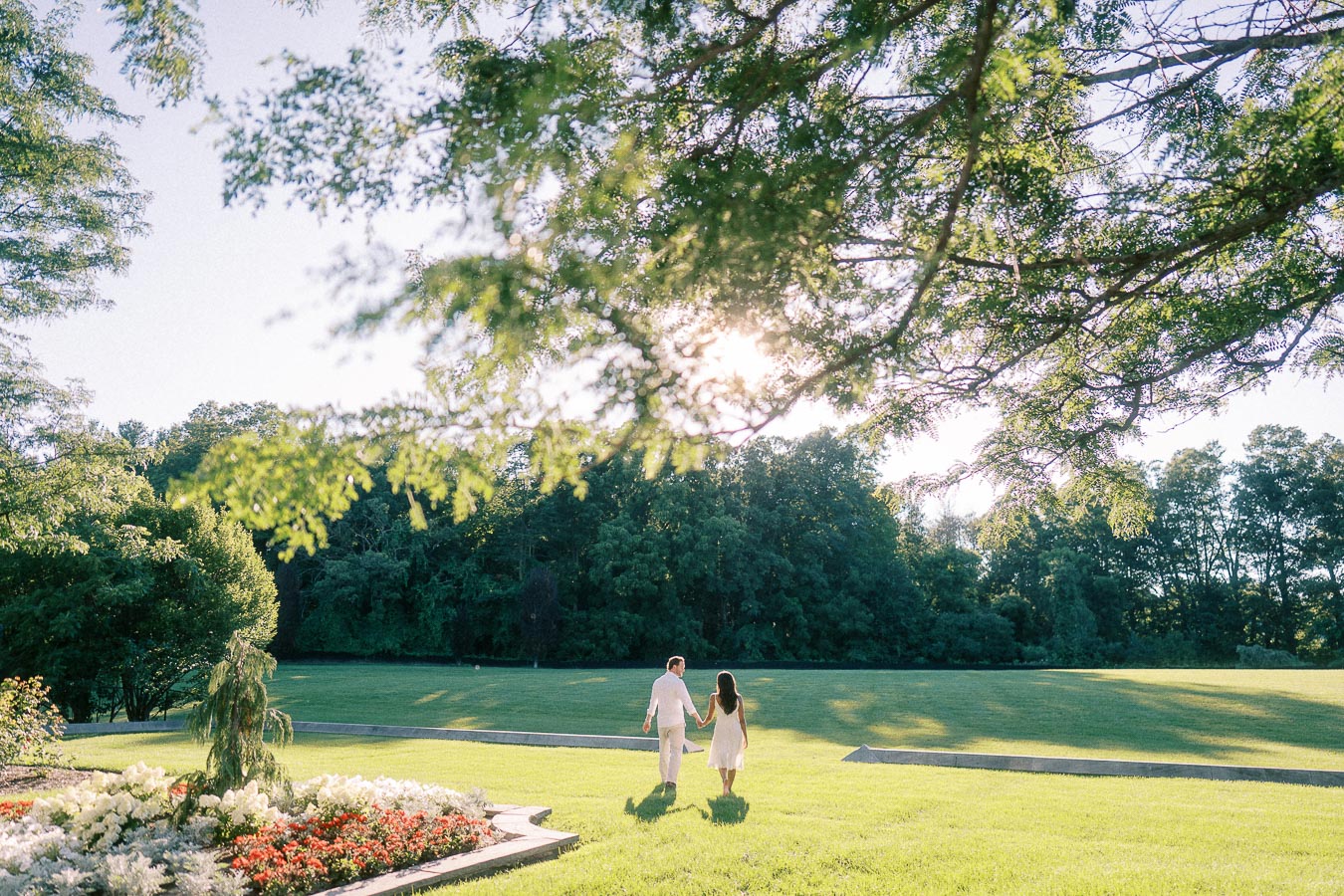 Rachel and Matt walk hand in hand across a sunlit lawn bordered by trees and garden flowers.