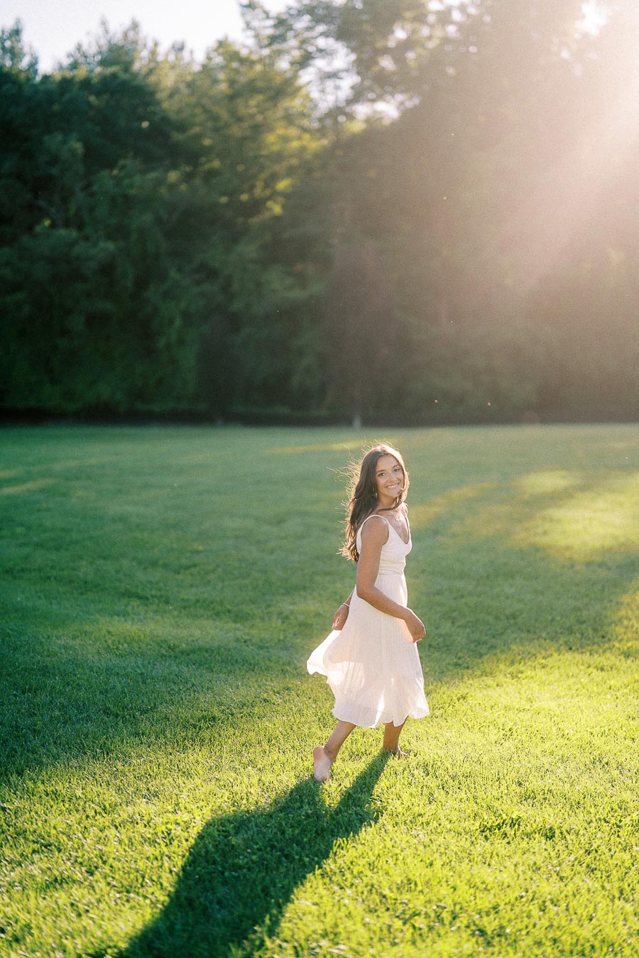 Rachel walks barefoot through a sunlit grassy field in a white dress, with trees behind her.
