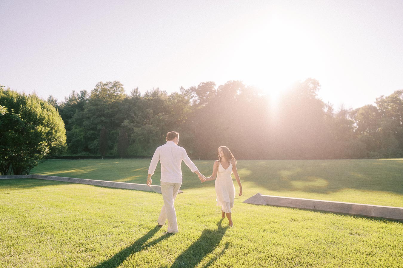 Rachel and Matt walk hand in hand across a sunny lawn with trees in the background in Aurora, Ontario.