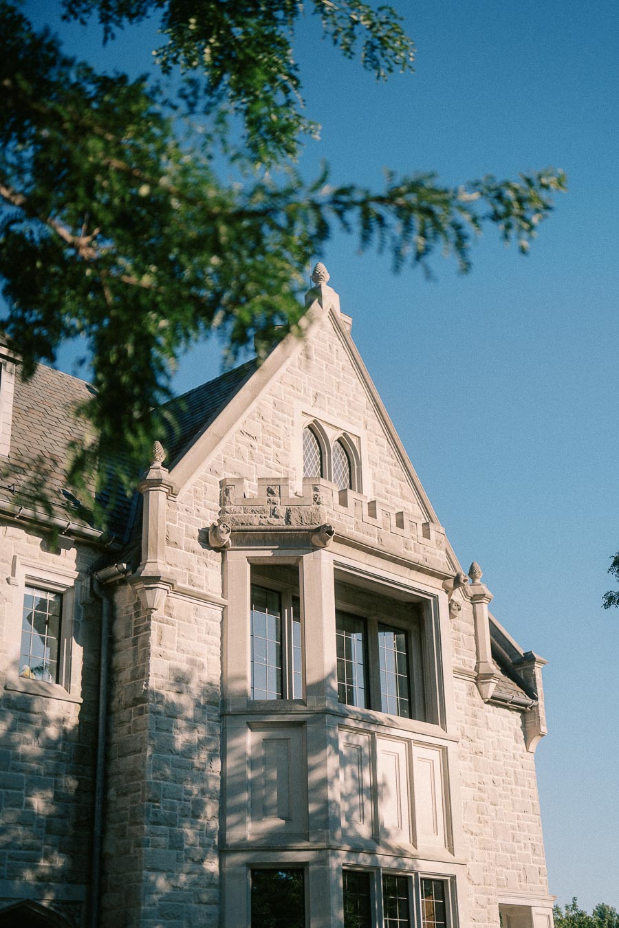 Sunlit stone school building with steep gables and leaded windows framed by leafy branches.