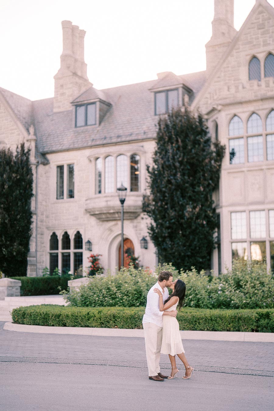 Rachel and Matt kiss in front of a stone Gothic-style building with tall windows and greenery.