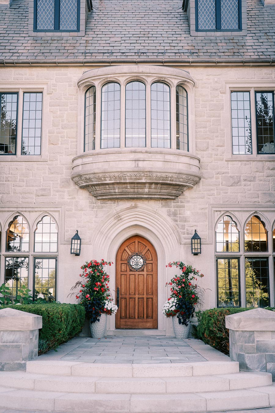 Stone school building entrance with arched wood door, bay window, leaded glass, planters, and steps.