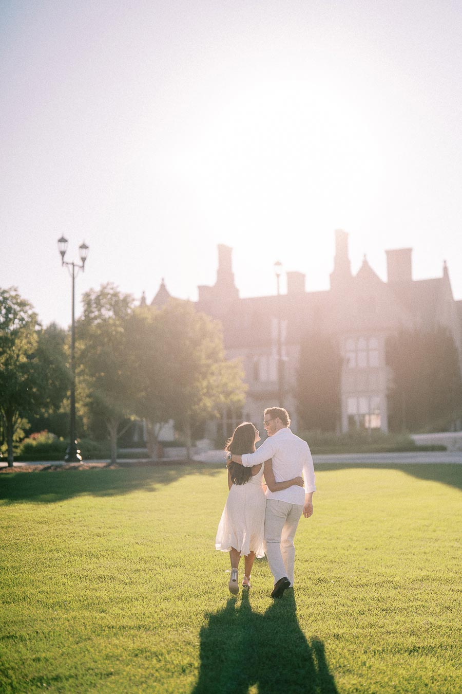 Rachel and Matt walk arm in arm across a sunlit lawn toward a historic school building in Aurora.