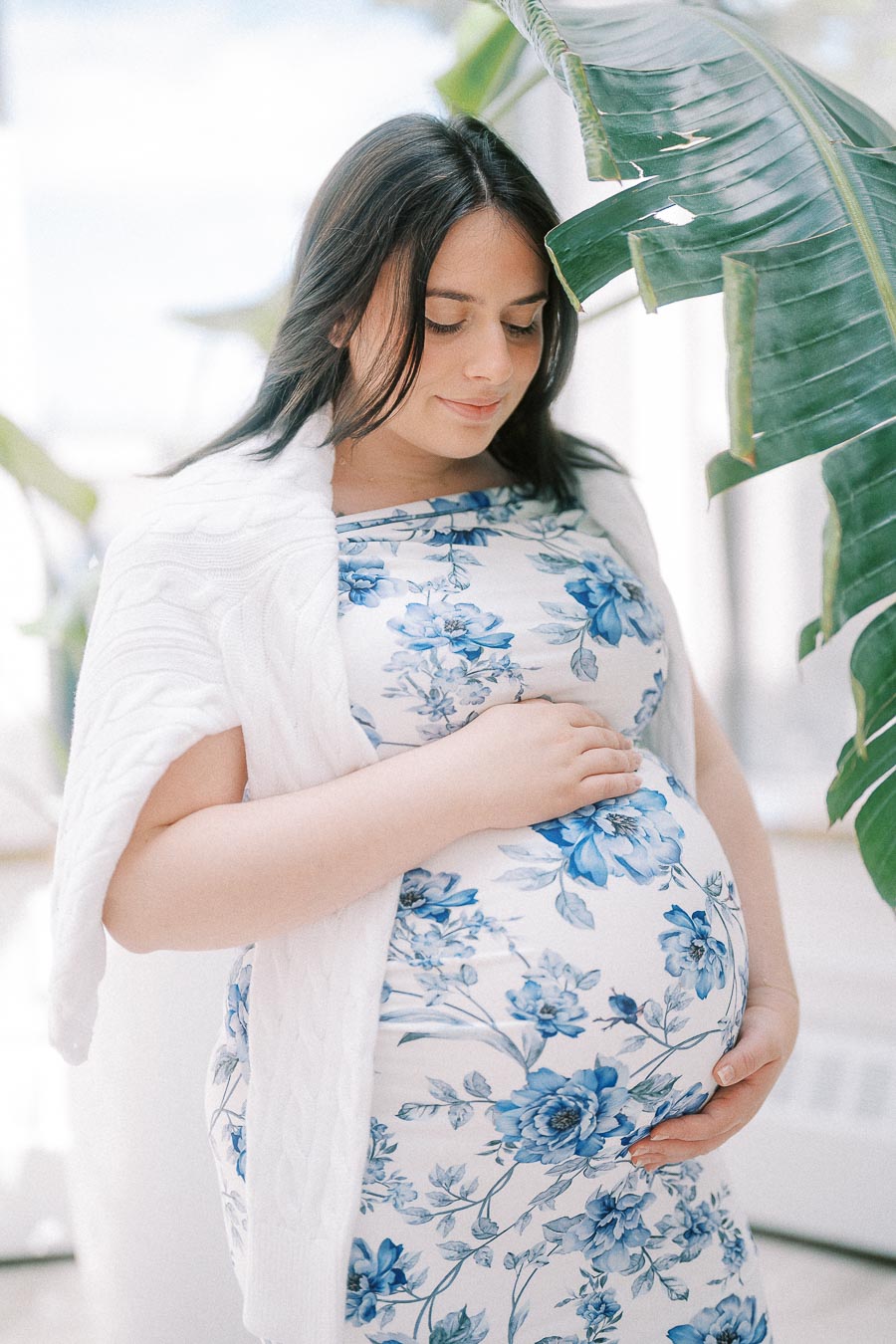 Pregnant woman in blue floral dress cradling her belly beside tropical leaves