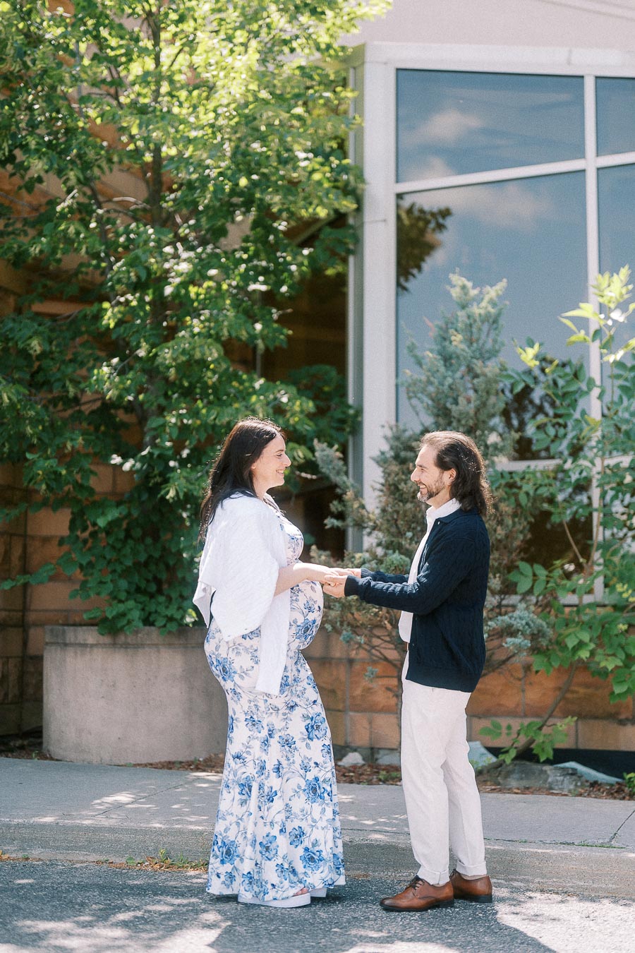 Pregnant couple holding hands outside a leafy building entrance.