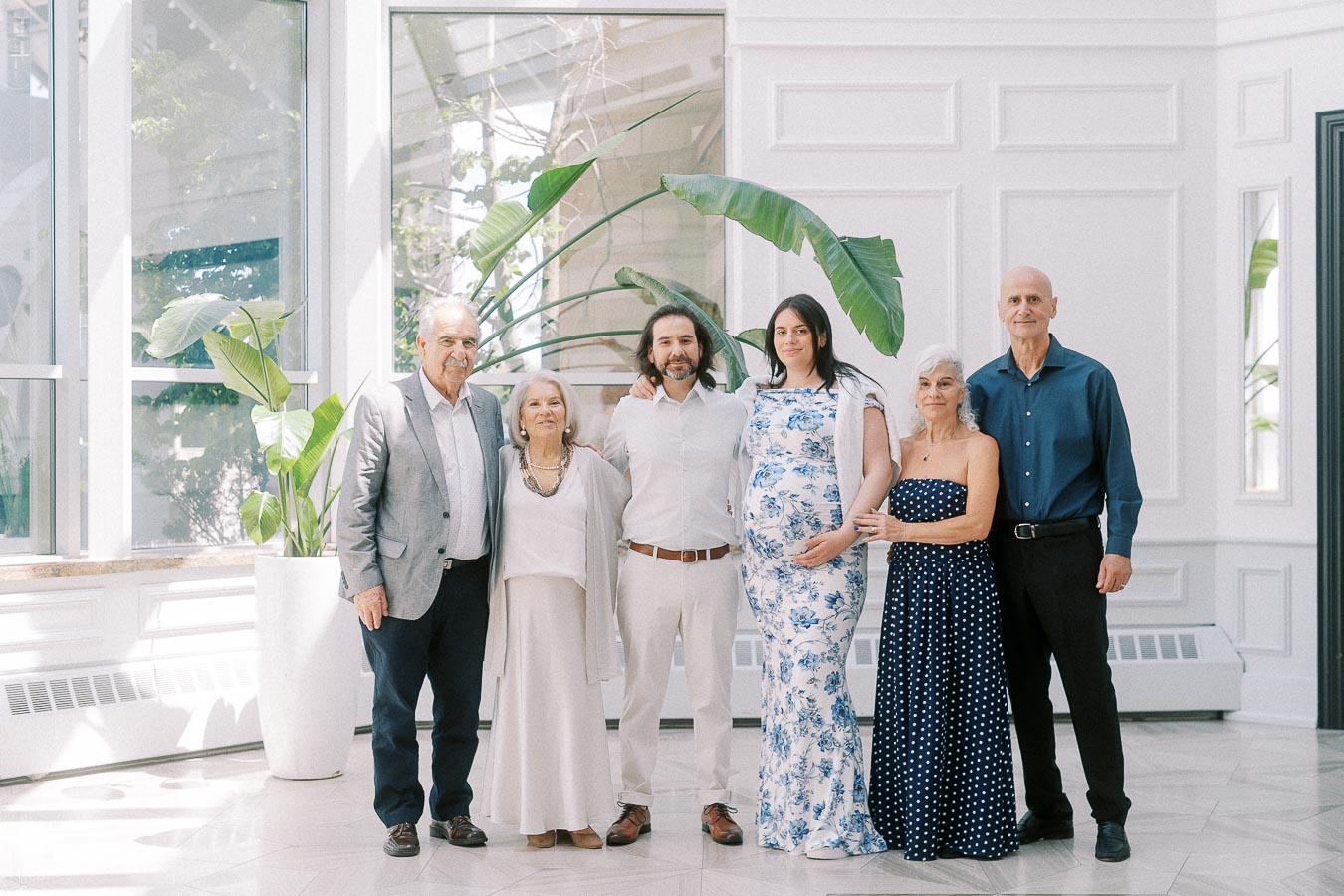 Six adults posing together in a bright room with tall plants.