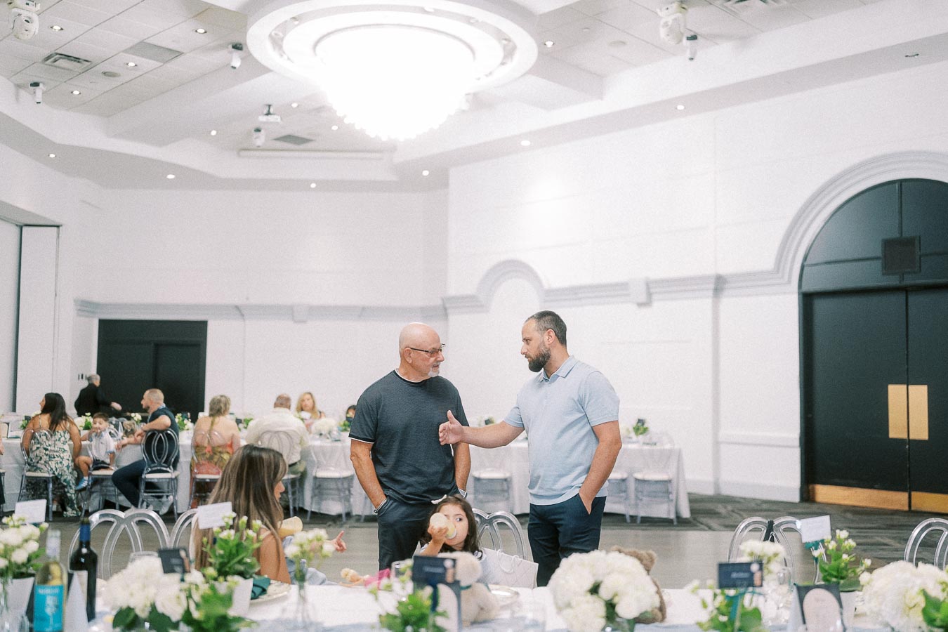 Two men talking among decorated tables in a bright event hall