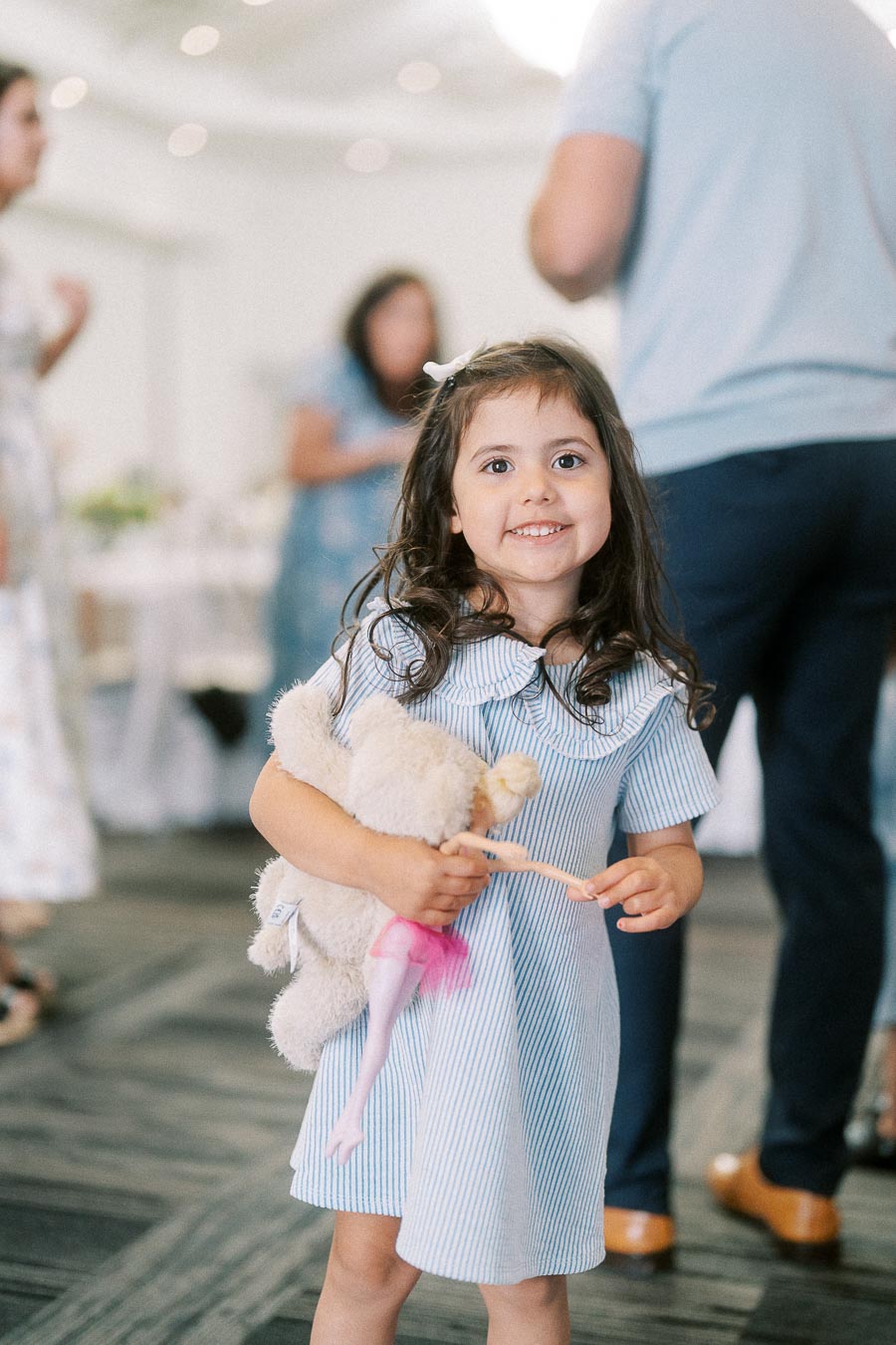 Smiling young girl holding a teddy bear and doll at an indoor celebration