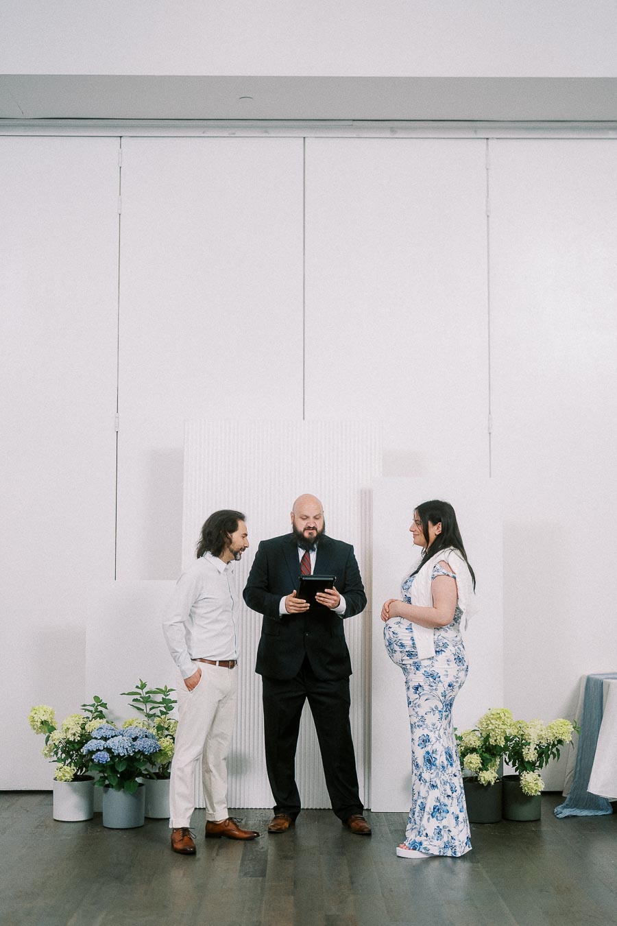 Couple stands with officiant before white backdrop and floral arrangements.