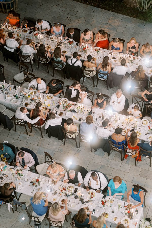 Aerial view of a formal outdoor banquet with elegantly dressed guests seated at long tables adorned with floral centerpieces, enjoying a festive meal under warm lighting.