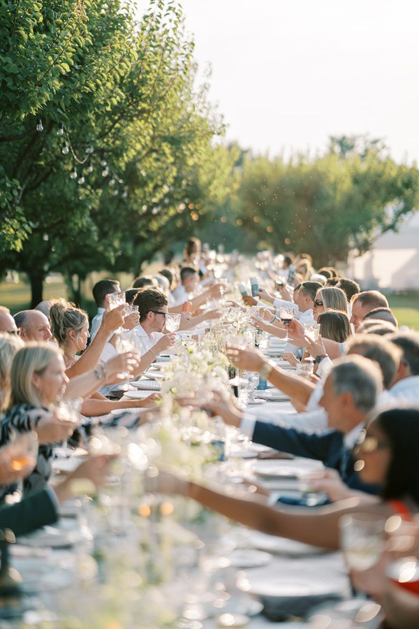 Outdoor wedding reception with guests raising glasses in a celebratory toast, seated at a long elegantly decorated table surrounded by lush greenery.