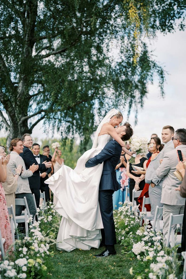 Bride and groom celebrating outdoors, embraced amidst cheering guests at a wedding ceremony, surrounded by white floral decorations and lush greenery.