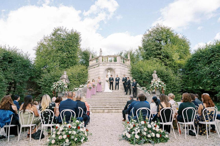 Elegant outdoor wedding ceremony set in a lush garden with a vintage stone staircase, surrounded by guests seated on ornate chairs, floral arrangements, and a bridal party standing on the steps against a backdrop of greenery and blue sky.
