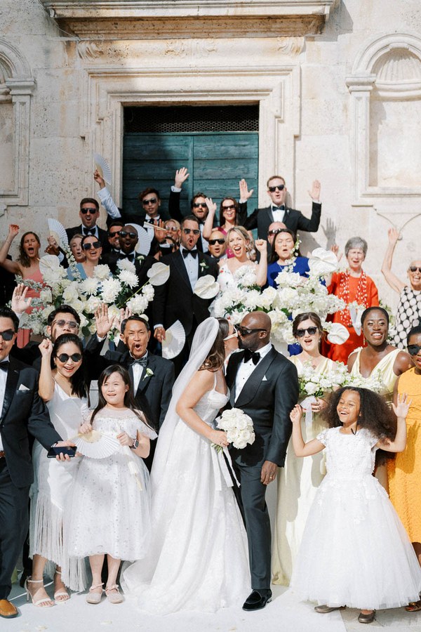 A joyful and diverse wedding group celebrating outside, featuring a bride in a white gown and veil kissing her groom in a tuxedo. They are surrounded by well-dressed guests holding white flowers, all smiling and raising hands in celebration in front of an ornate building.