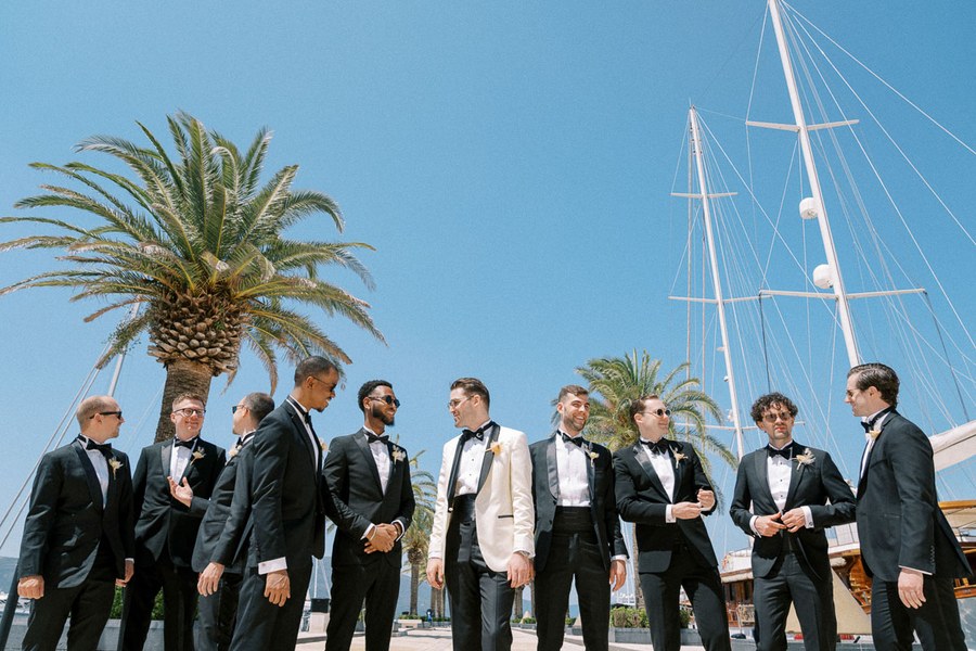 Groom and groomsmen in tuxedos at an outdoor wedding by a marina, featuring palm trees and a clear blue sky.