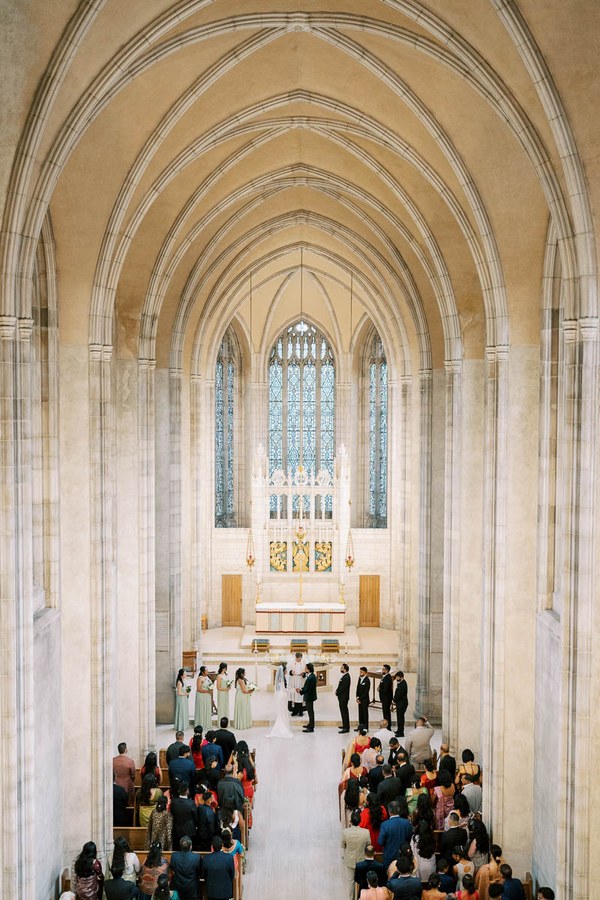 A beautifully constructed cathedral with an ornate, vaulted ceiling hosting a wedding ceremony. The bride and groom stand at the altar with bridesmaids and groomsmen in attendance, as guests fill the pews. The stained glass windows and intricate designs add elegance to the spiritual atmosphere.