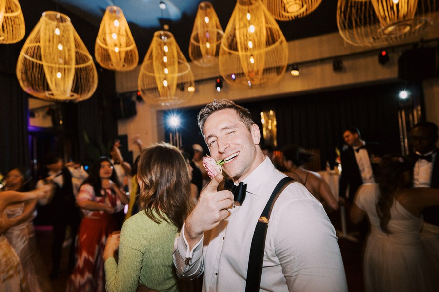 Man in formal attire playfully holding a flower with his mouth at a lively wedding reception, surrounded by dancing guests and warm lighting.