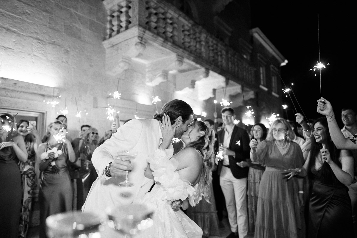 Wedding photography questions about candid moments as a bride and groom kiss during a sparkler exit surrounded by celebrating guests