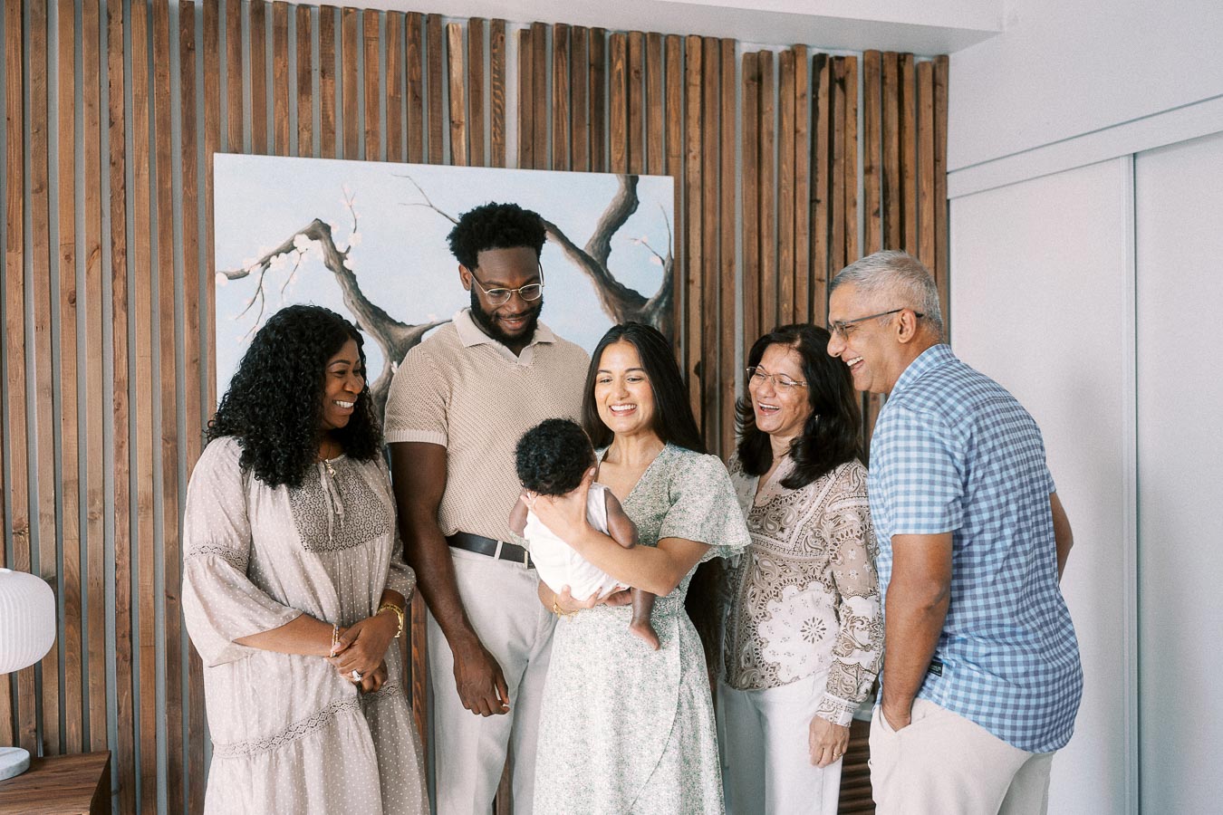 Family members gather around Karina holding Isabella beside a wood slat wall.
