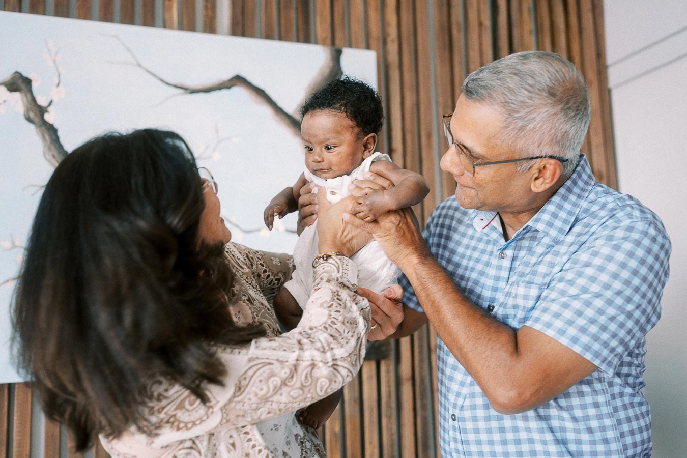 Karina and Terence holding baby Isabella beside a wood slat wall and abstract artwork.