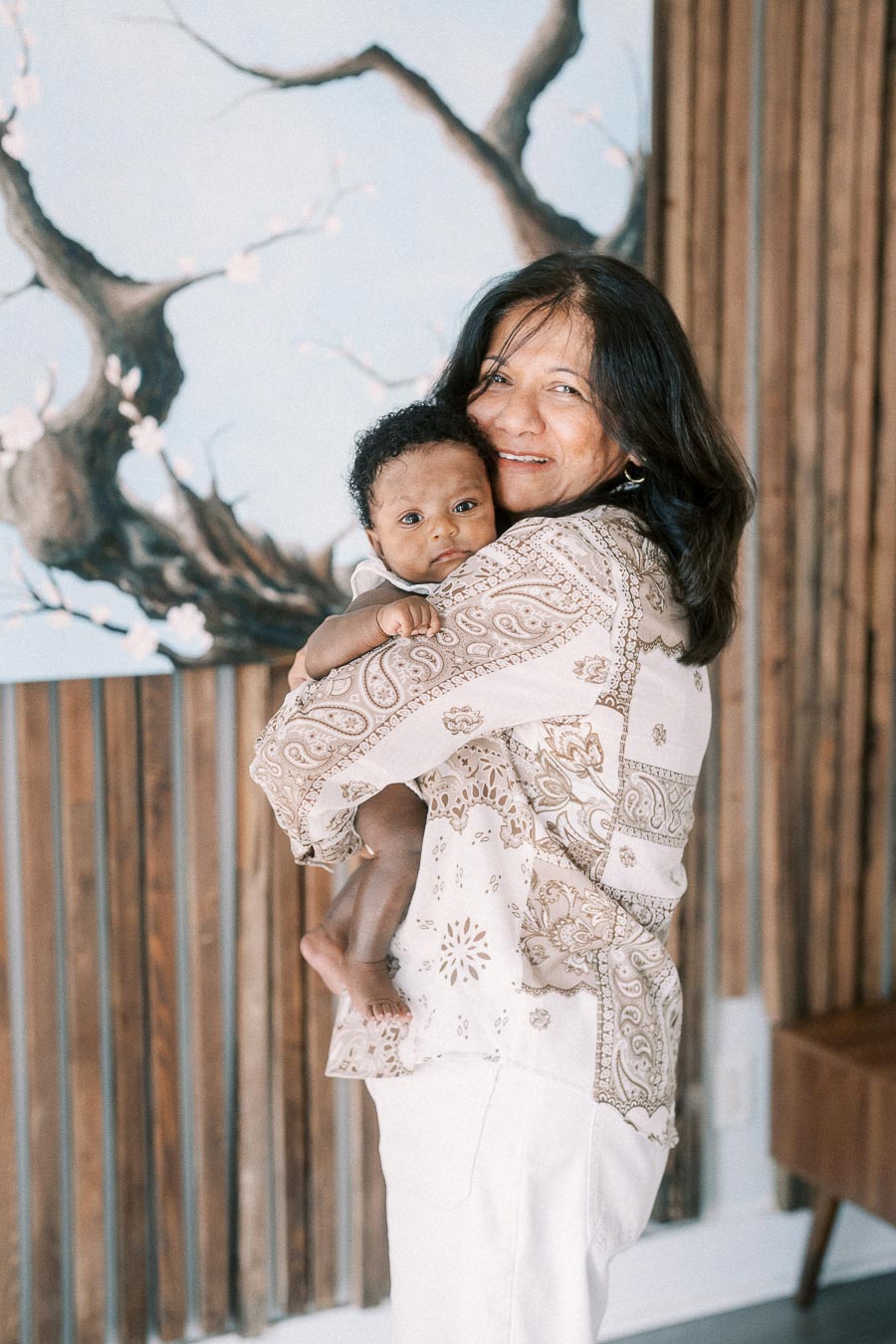 Karina smiles while holding Isabella against a wood-paneled wall and tree artwork.