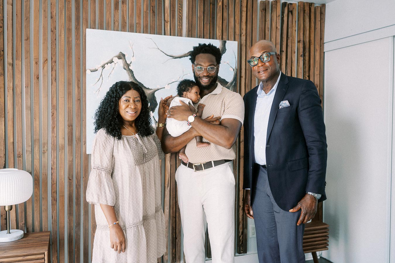 Family members stand with a newborn held by her father beside a wooden slat wall.