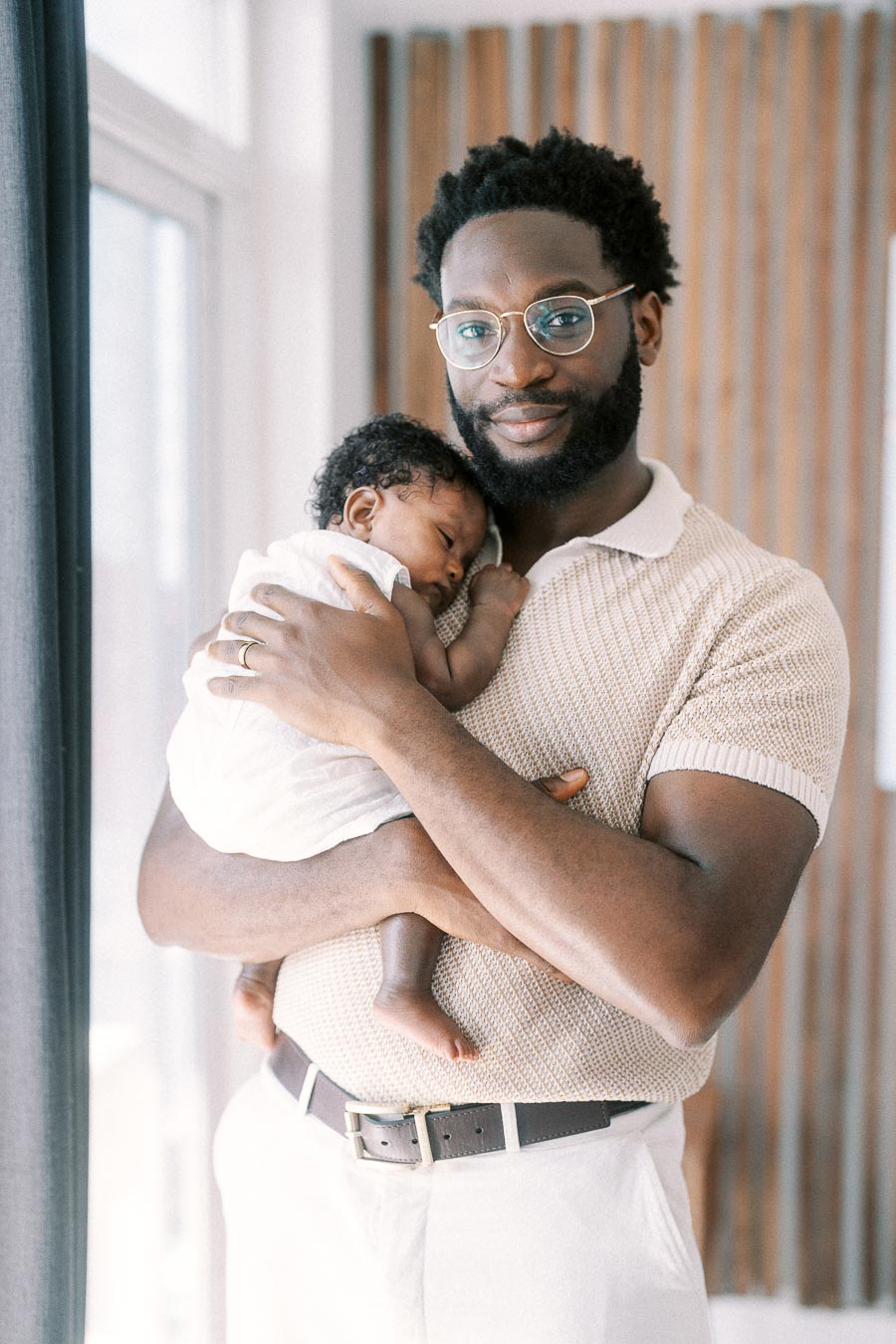 Terence holds sleeping newborn Isabella against his chest beside a bright window.