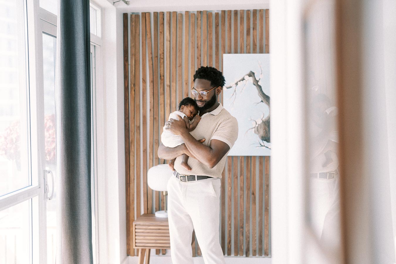 Terence cradles newborn Isabella beside a bright window and wood slat wall.