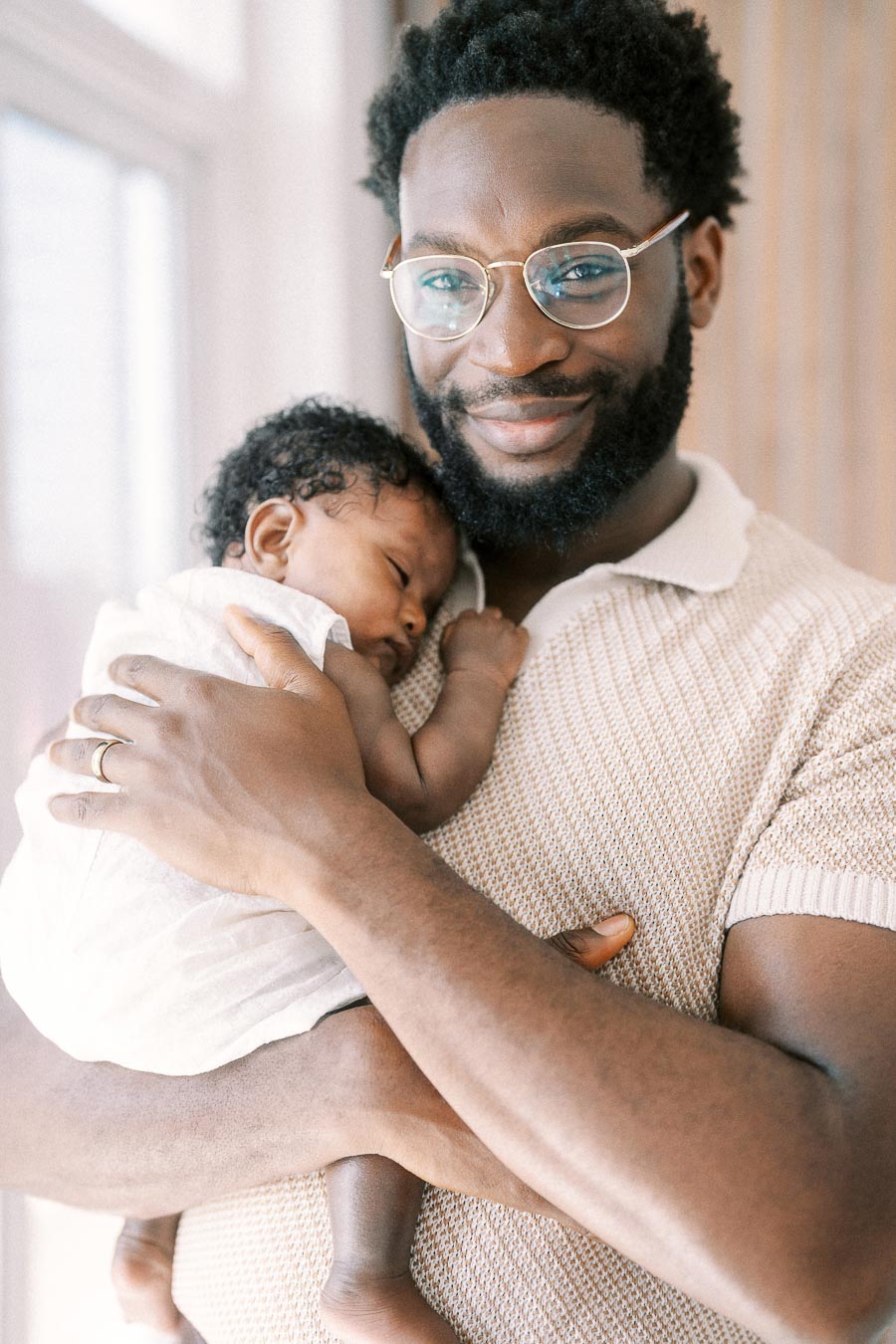 Terence cradles sleeping newborn Isabella against his chest by a bright window.