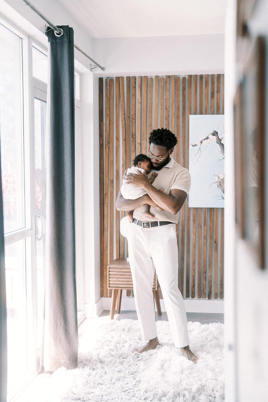 Terence cradles newborn Isabella beside a bright window and wood slat wall.