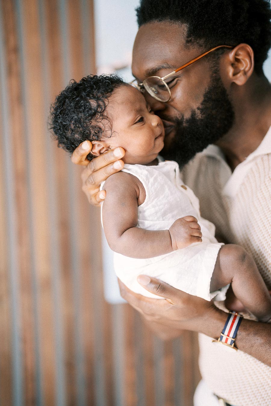 Terence kisses newborn Isabella while holding her in a white outfit at home.