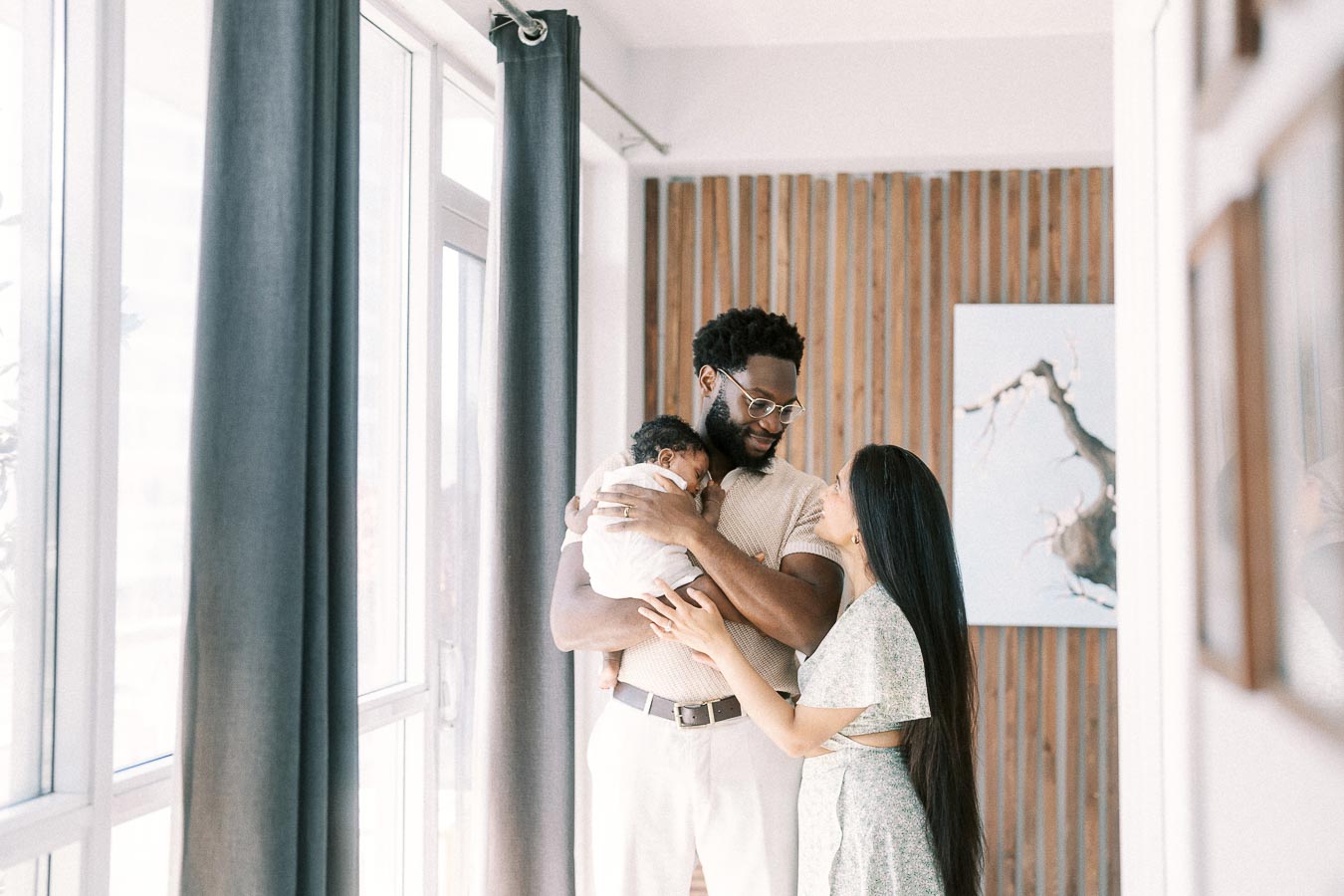 Karina and Terence cuddle newborn Isabella beside tall windows in their Toronto home.