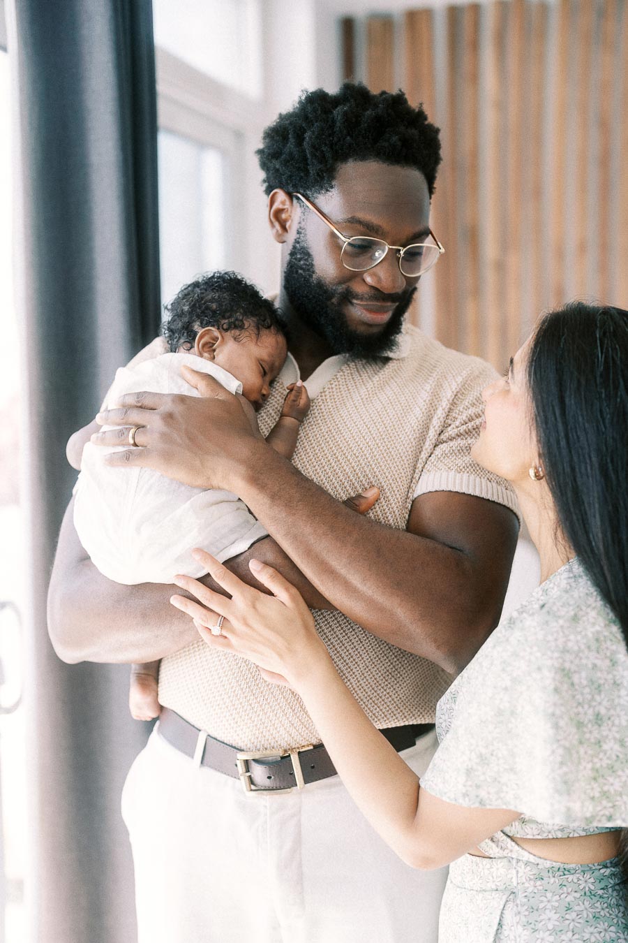 Father holding newborn daughter as mother smiles beside them near a bright window.