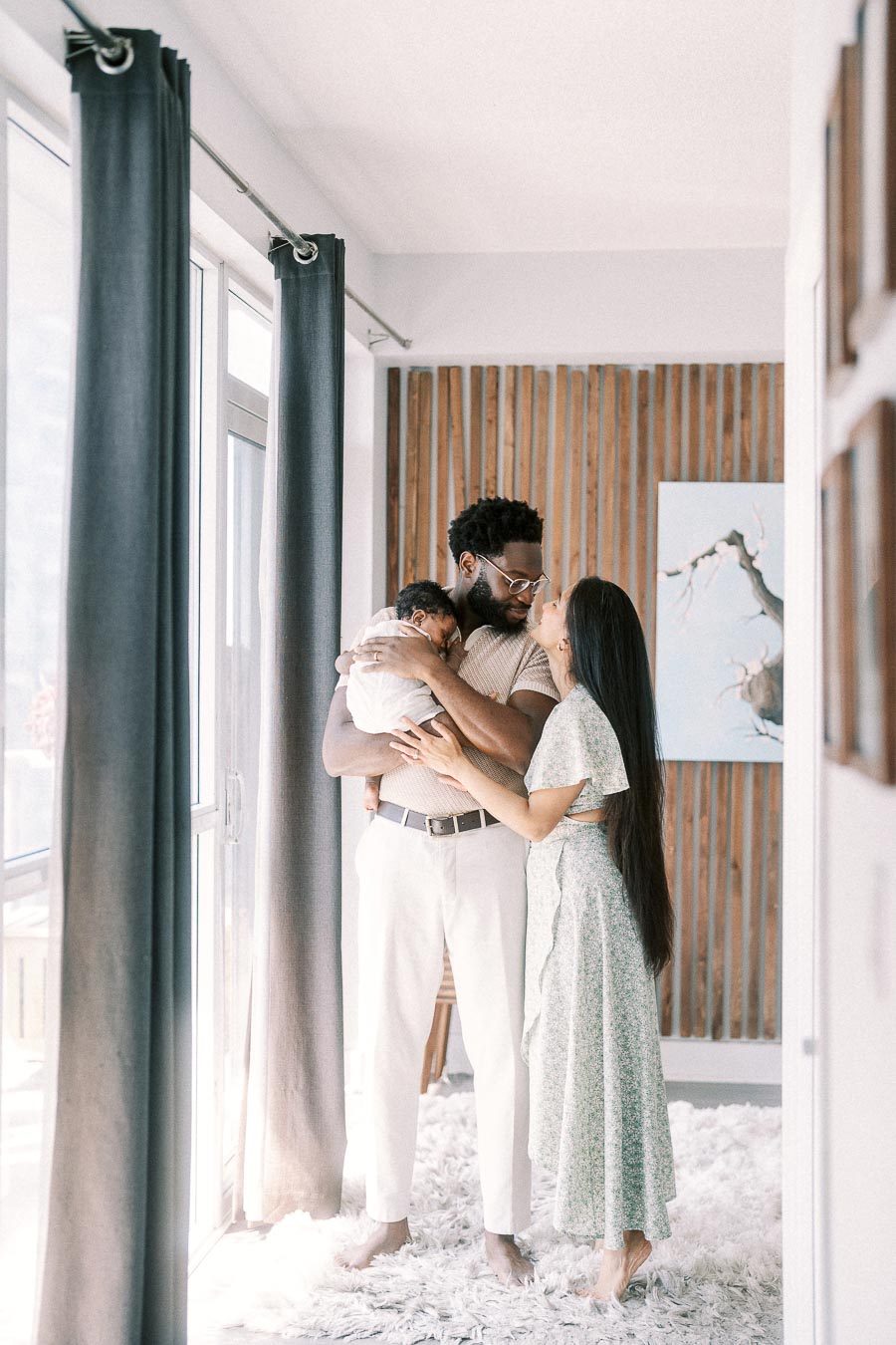 Parents cuddle newborn daughter by tall windows during an in-home newborn photography in Toronto session.