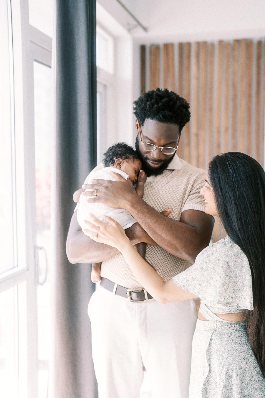 Terence holds newborn Isabella as Karina gently supports her beside a bright window.
