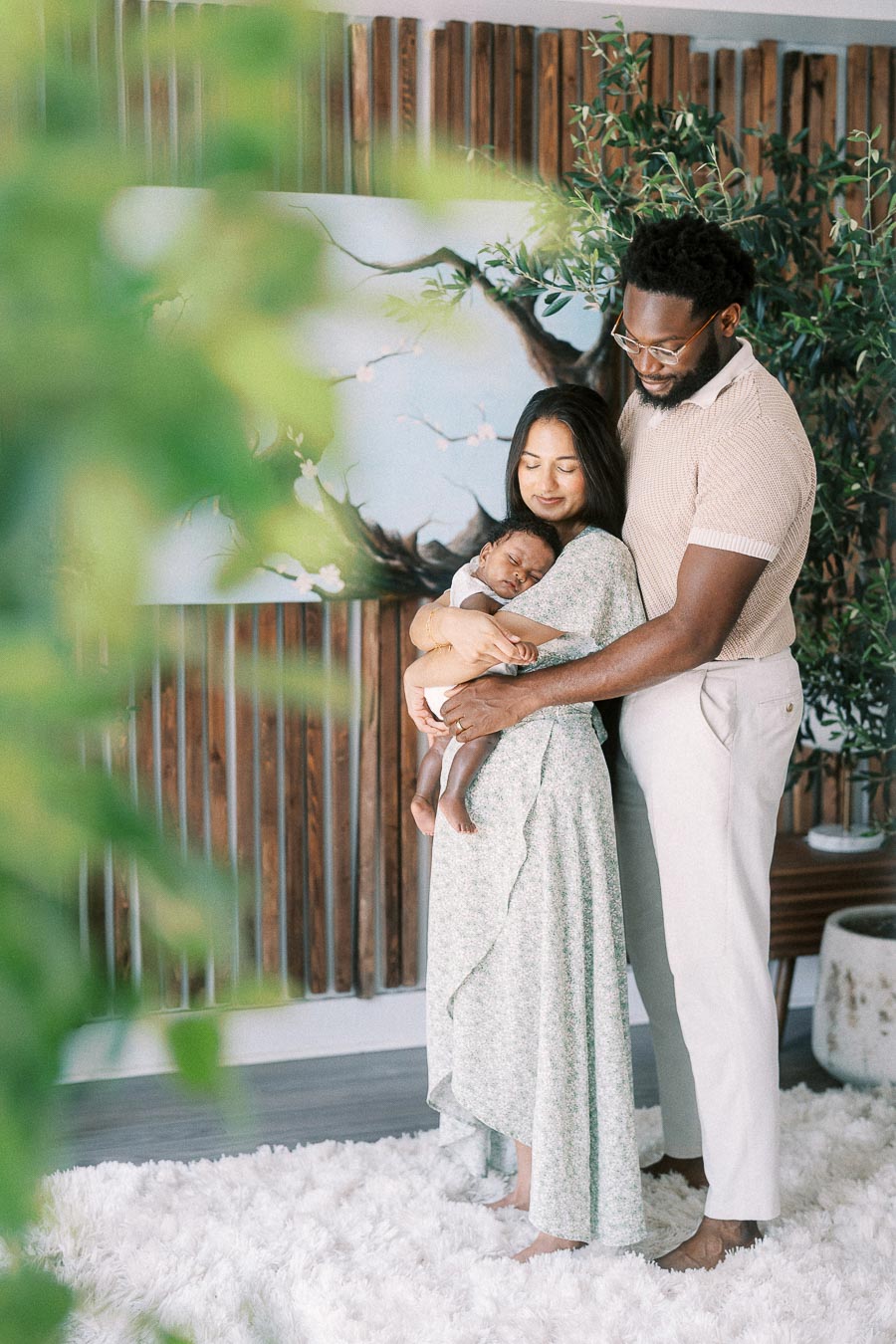 Parents holding their newborn daughter beside indoor greenery and a wood accent wall.
