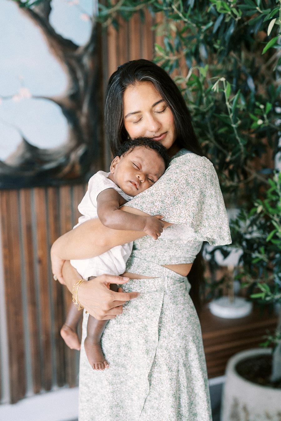 Karina cradles sleeping Isabella beside leafy plants in their Toronto home.