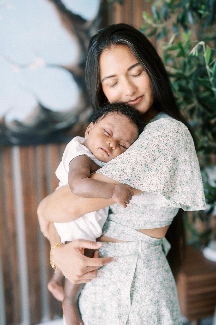 Karina cuddles sleeping newborn Isabella against her shoulder beside indoor greenery.