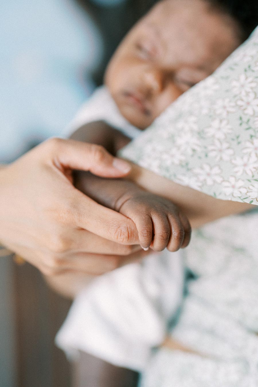 Newborn Isabella holds an adult finger while sleeping in a floral blanket.
