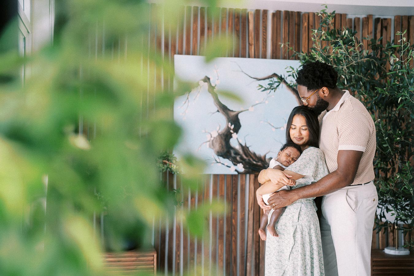 Karina and Terence cuddle newborn Isabella beside plants and wood panelling at home.